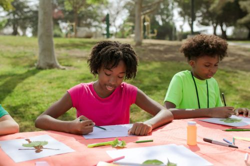 Two children engage in creative drawing activities outdoors in a sunny park setting.